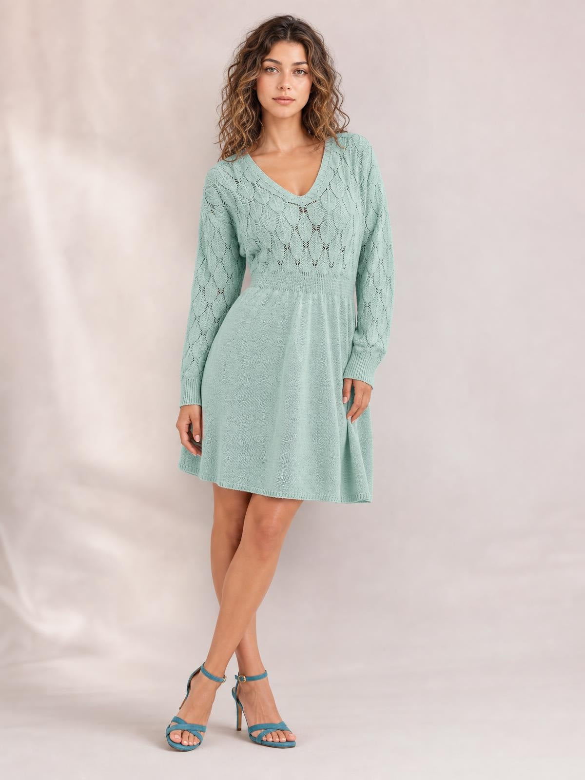 A model poses with crossed legs in a mint green V-neck sweater dress featuring long sleeves, openwork knit detailing, a fitted waist, and a flared silhouette.