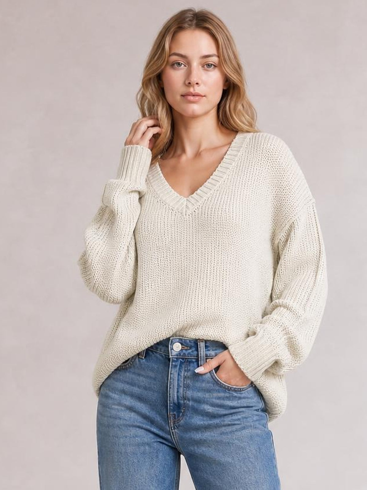 Model poses in a cream V-neck loose knit drop shoulder sweater with ribbed cuffs and neckline, relaxed oversized silhouette, and exposed seams.