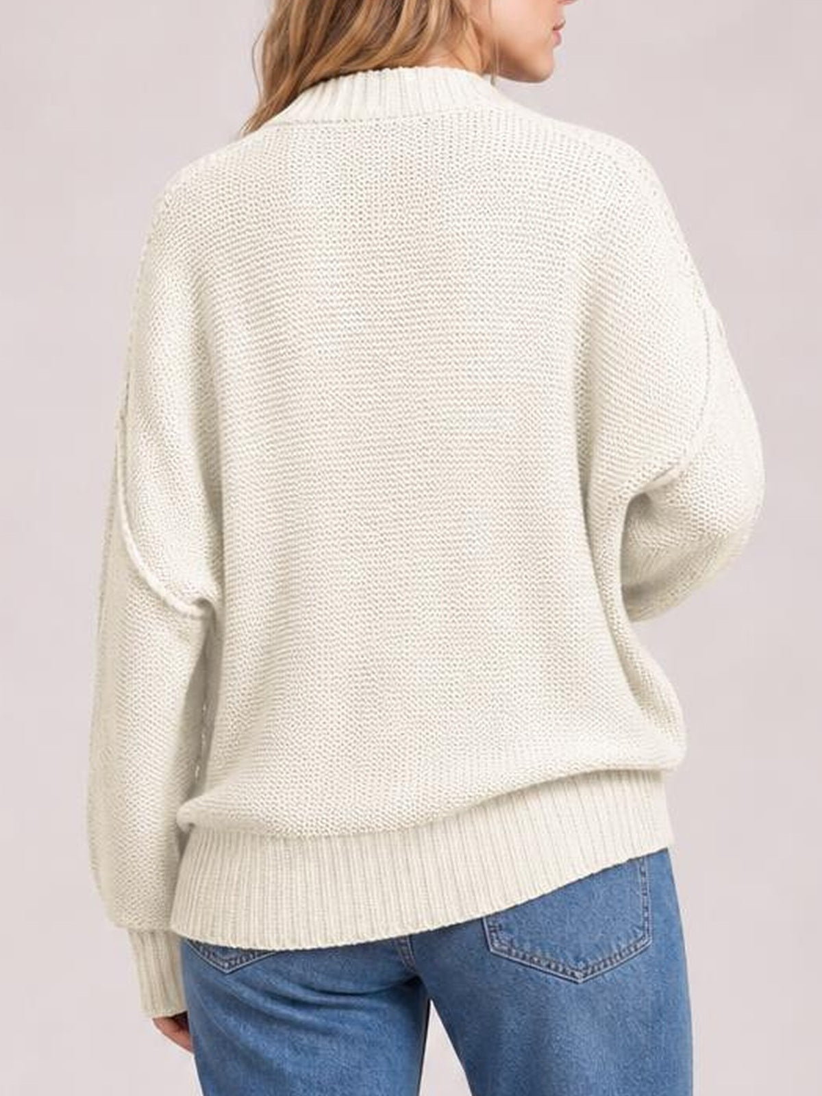 Model turned away wears a cream loose knit drop-shoulder sweater with an oversized silhouette, ribbed cuffs and hem, and exposed seam detailing.