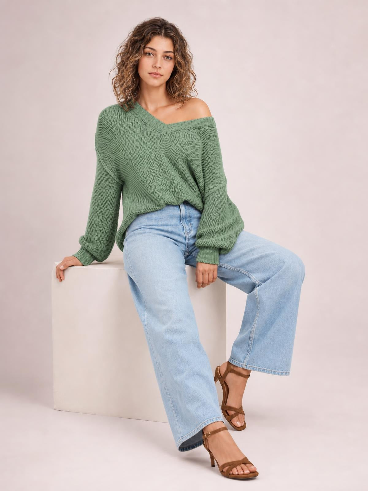 Model seated in a sage green V-neck loose knit sweater with drop shoulders, oversized silhouette, ribbed cuffs, and visible exposed seam detailing.
