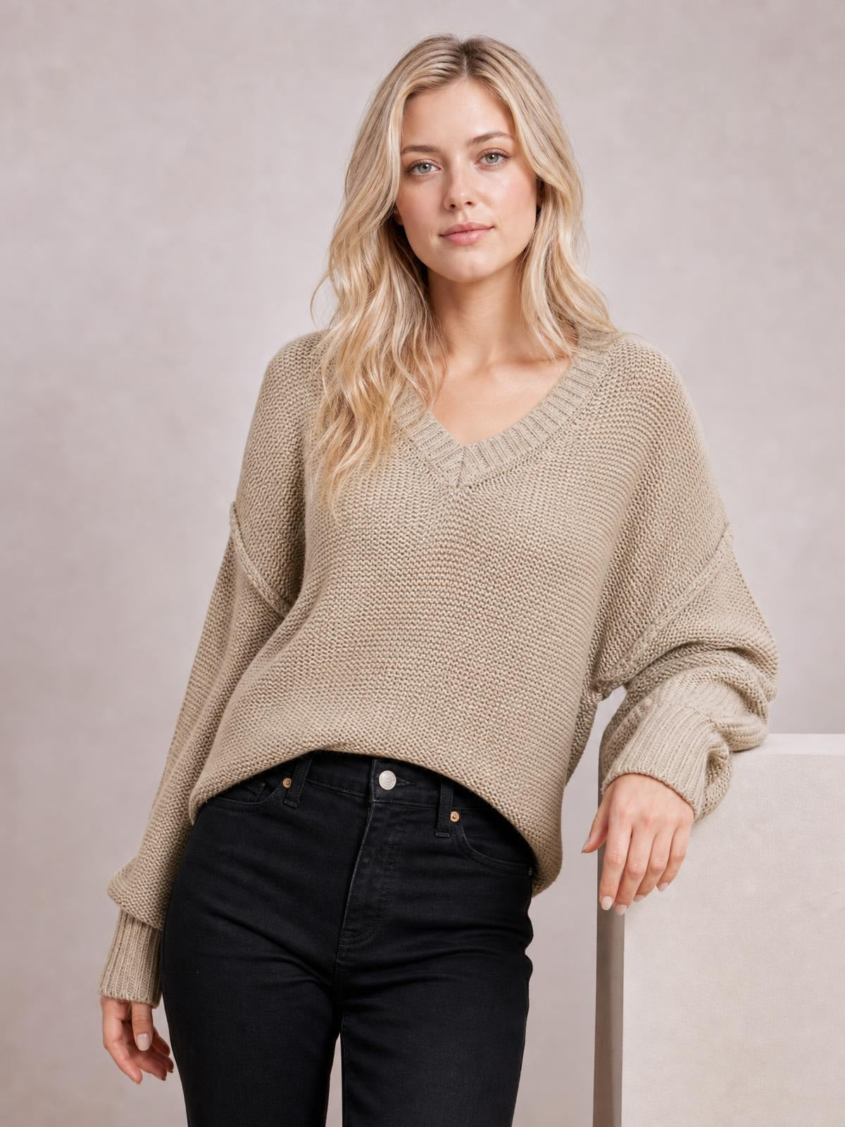 Model wearing a taupe V-neck loose knit sweater with a relaxed drop-shoulder silhouette, ribbed trim, exposed seams, and long sleeves.