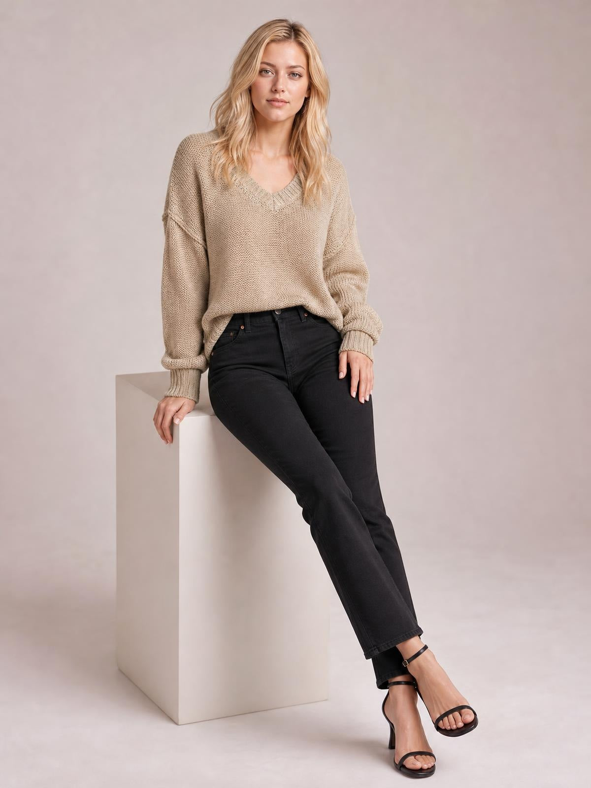 Model seated in a taupe V-neck loose knit sweater with drop shoulders, exposed seams, ribbed trim, relaxed silhouette, and rolled cuffs.