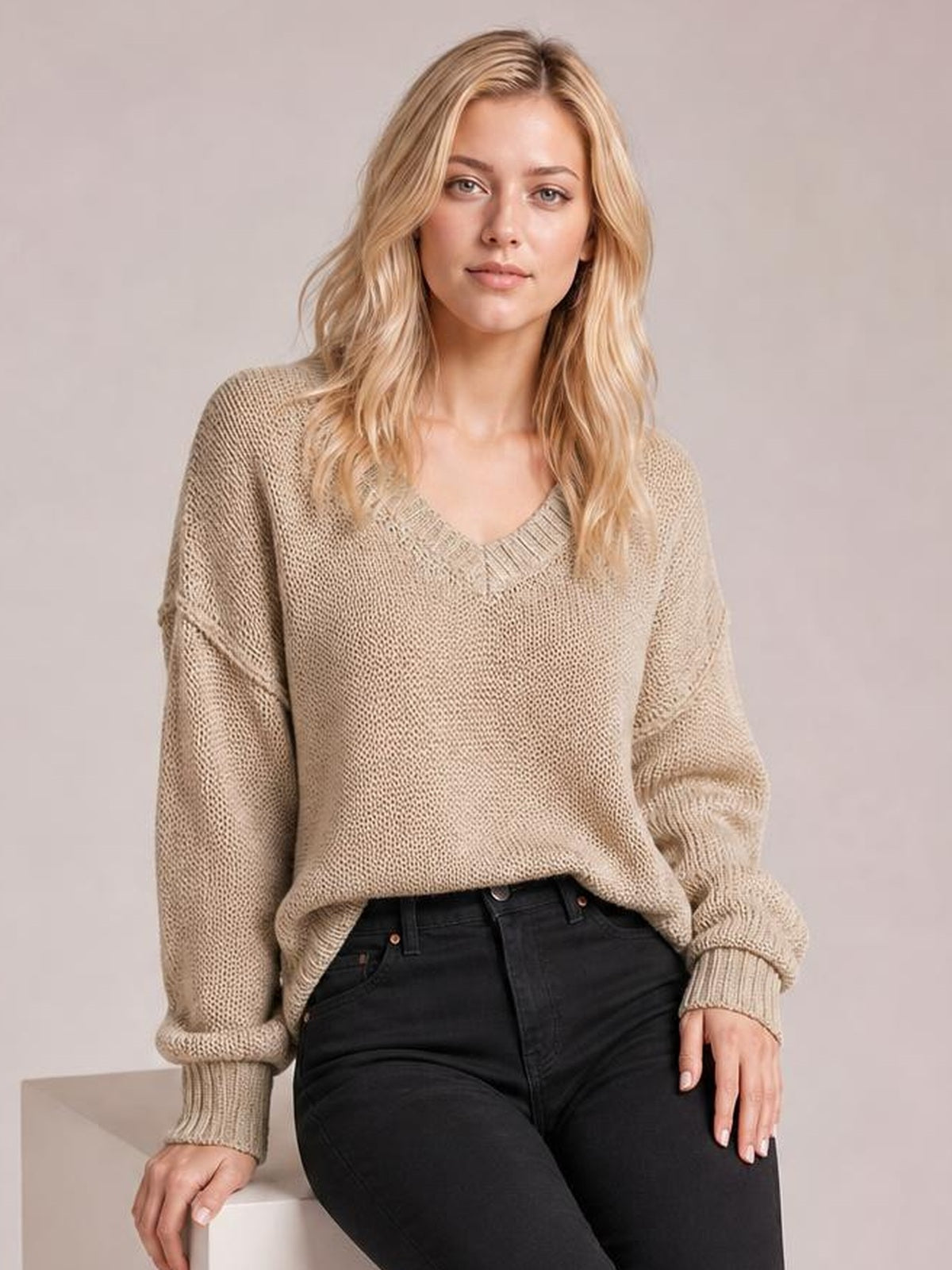 Model seated in a beige loose-knit V-neck sweater with drop shoulders, ribbed trim, exposed seams, and a relaxed oversized silhouette.