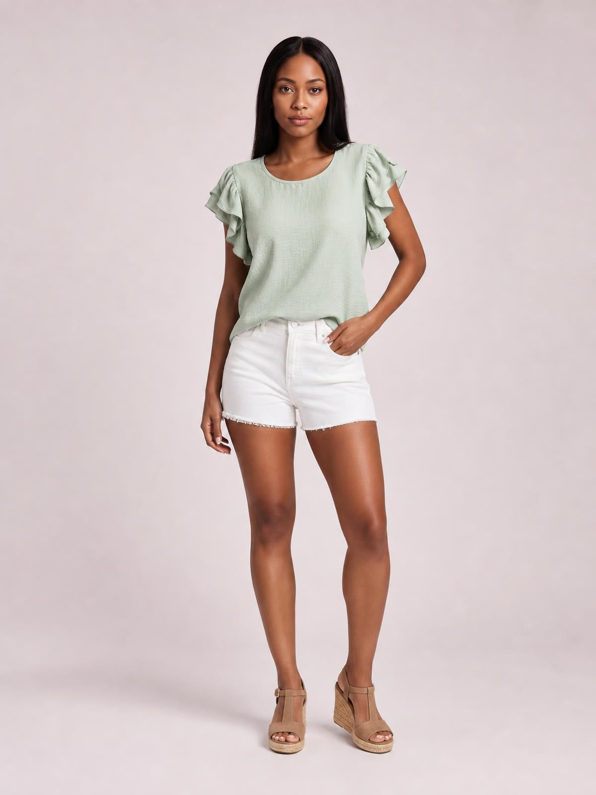 Model wearing a sage green blouse with layered ruffled cap sleeves, a rounded neckline, and a relaxed straight silhouette, standing with one hand in her pocket.