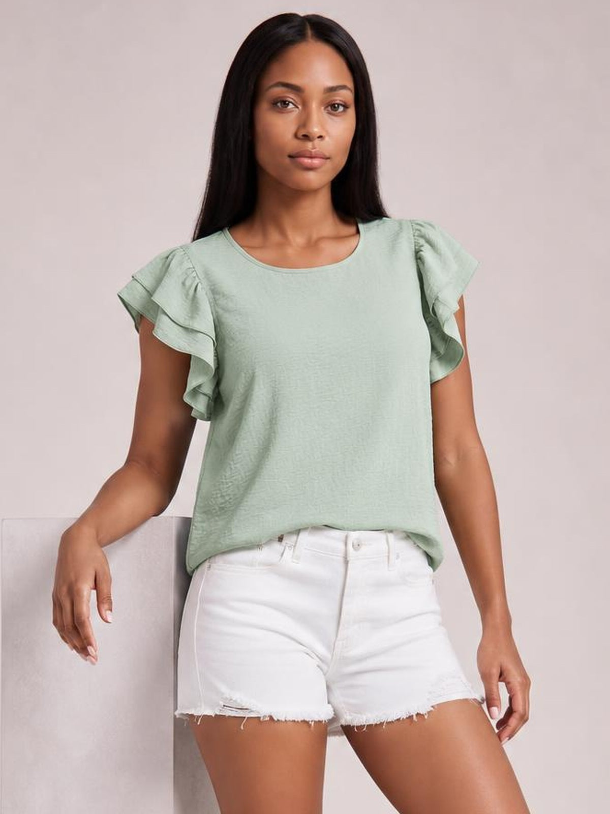 A model wears a mint green blouse with layered ruffled cap sleeves, a rounded neckline, and a relaxed straight silhouette, posed with one arm resting at her side.