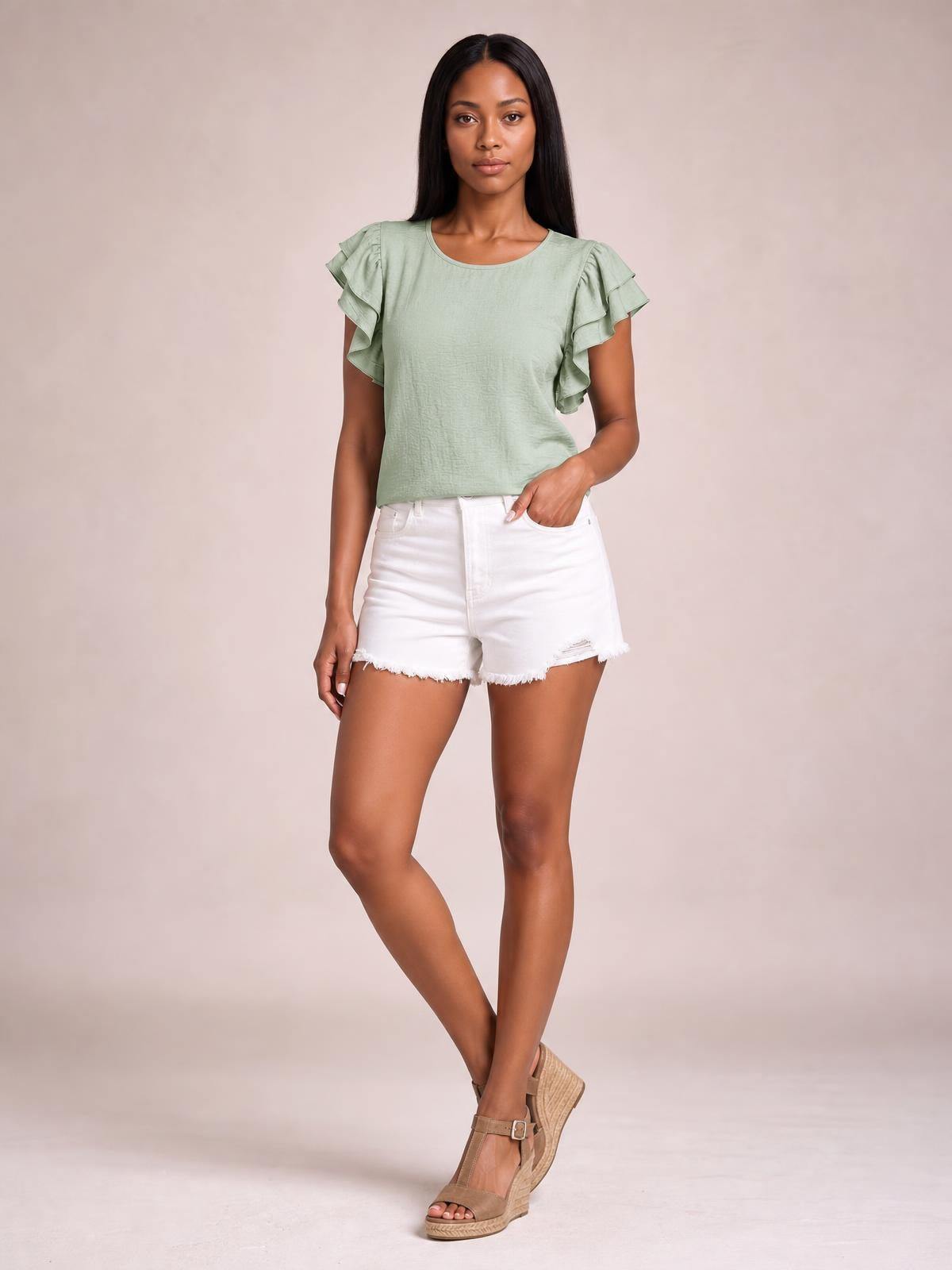 Model wears a sage green blouse with a round neckline, layered ruffled cap sleeves, and a relaxed straight silhouette, posing with one hand in her shorts pocket.
