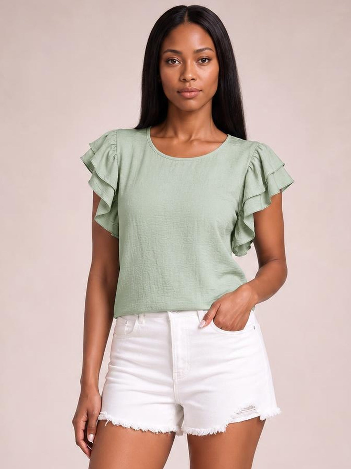 A model wears a sage green blouse with layered ruffled cap sleeves and a relaxed straight silhouette, standing with one hand in her shorts pocket.