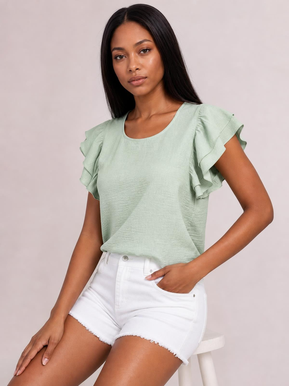 Model seated in a sage green blouse with a rounded neckline, layered ruffled cap sleeves, and a relaxed straight silhouette, one hand resting in her shorts pocket.