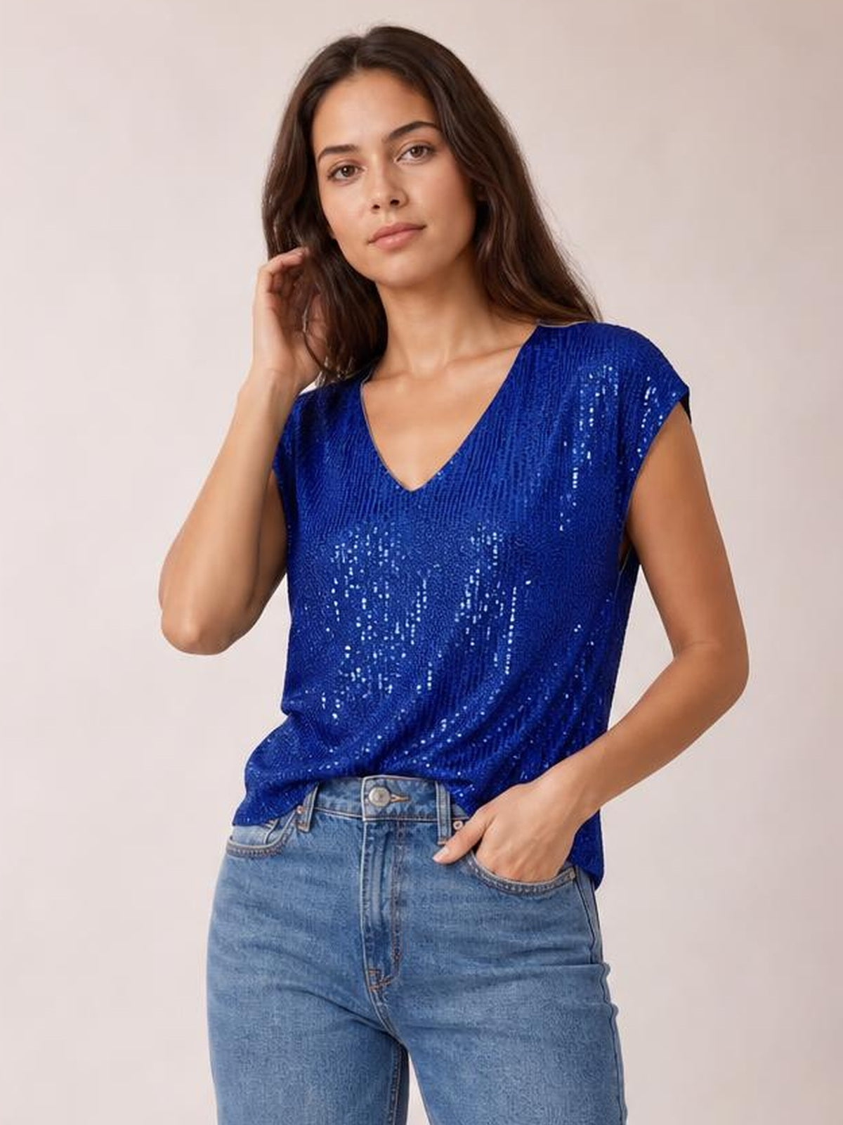 Model wearing a royal blue sequin top with a V-neck, cap sleeves, relaxed straight silhouette, and slightly draped hem, posing with one hand in her jeans pocket.