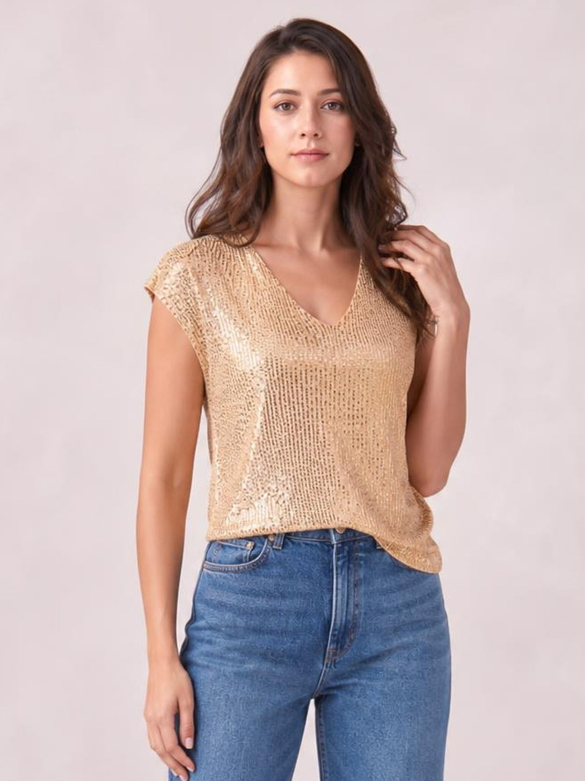 Model wearing a champagne-gold sequin V-neck top with cap sleeves and a relaxed straight silhouette, posing with one hand touching her hair.