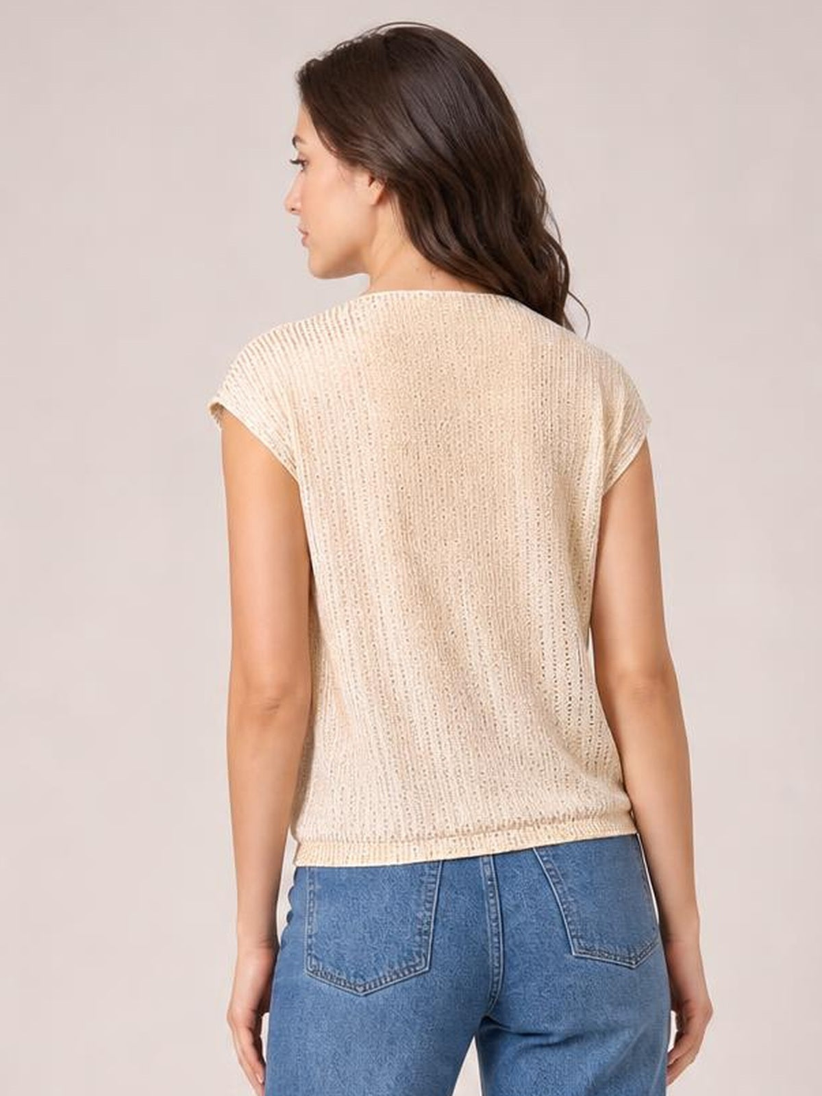 A model stands turned away wearing a cream sequin top with short cap sleeves, a relaxed straight silhouette, and a clean hem over blue jeans.