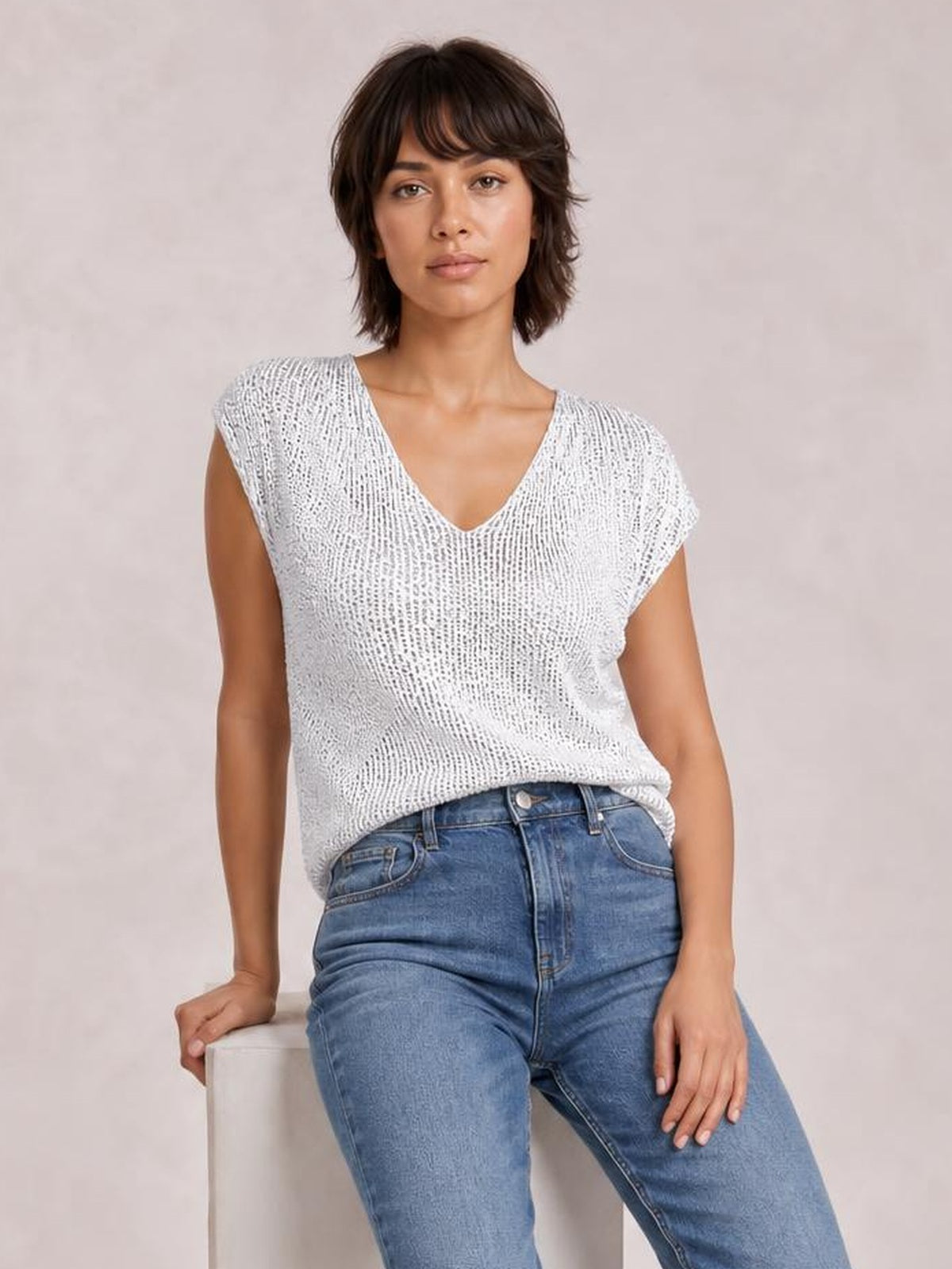 Model seated in a white sequin top with a V-neck, cap sleeves, relaxed straight silhouette, and slightly draped hem styled with blue jeans.