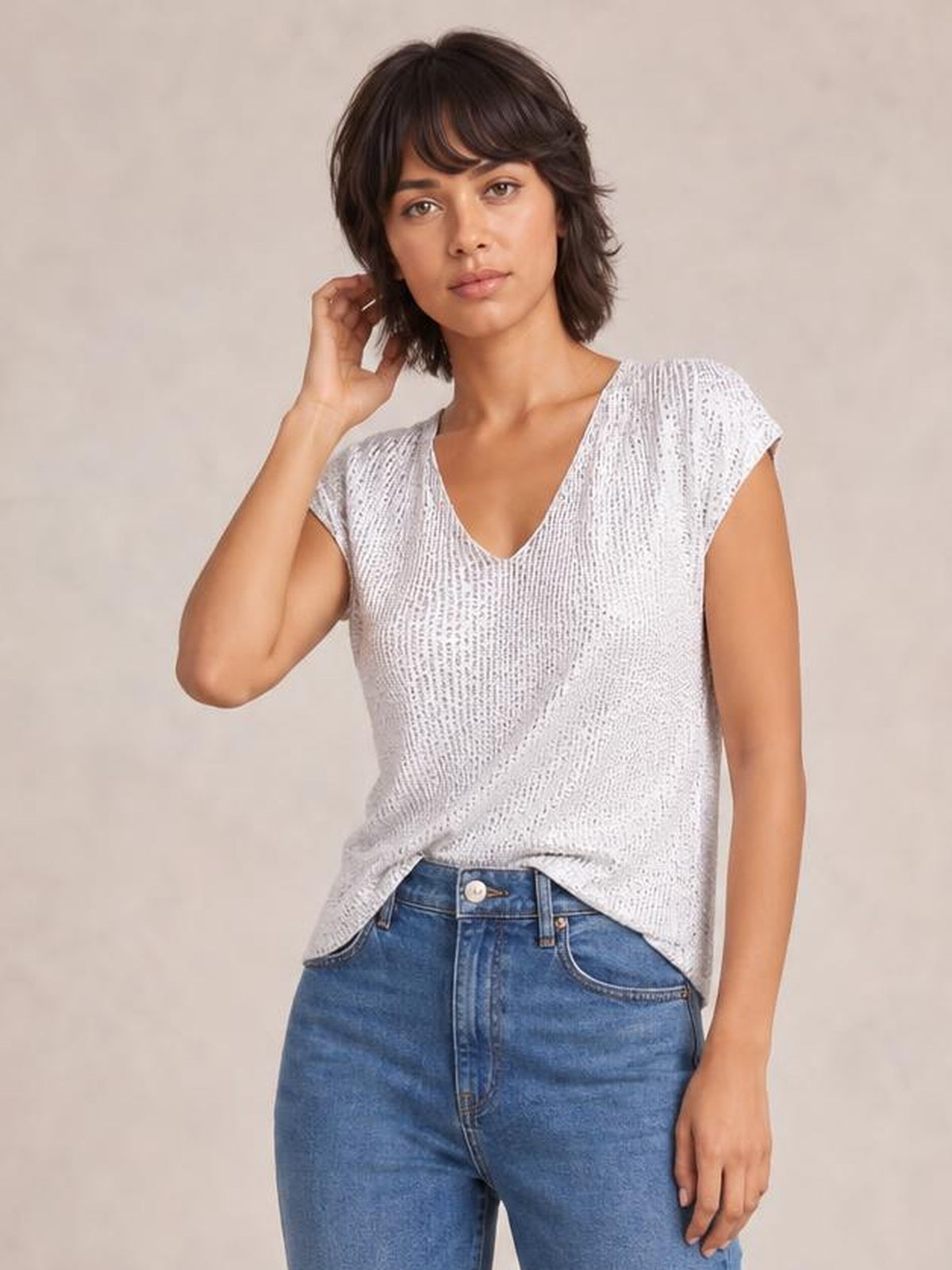Model wearing a white sequin V-neck cap sleeve top with a relaxed straight silhouette, paired with blue jeans, touching her hair in a casual pose.