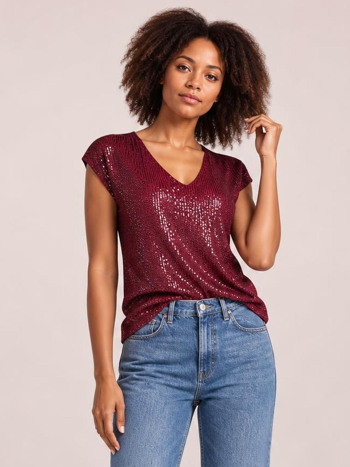 A model poses in a burgundy sequin top with a V-neck, short cap sleeves, and a relaxed silhouette, lifting one hand toward her hair.