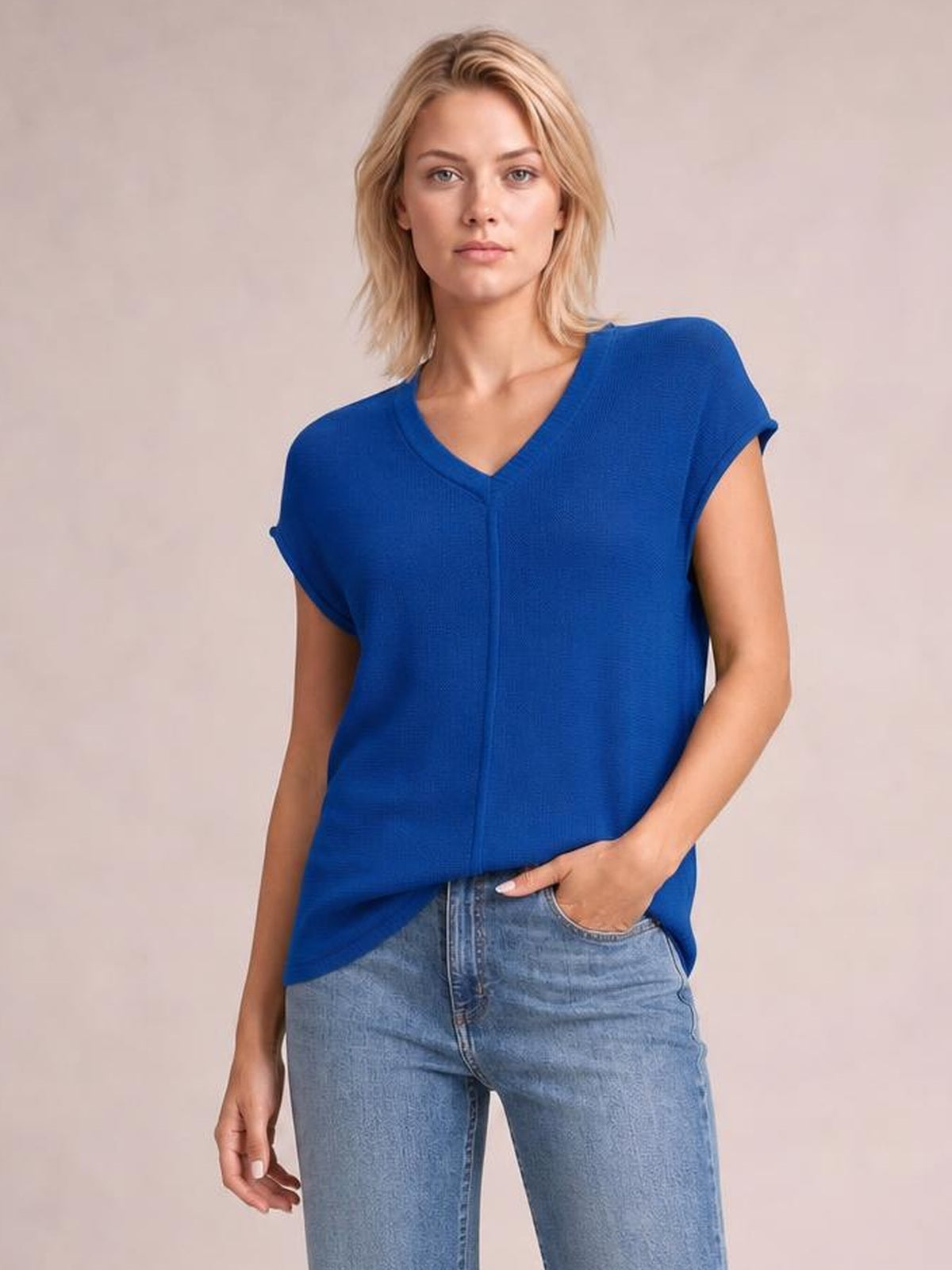 Model wearing a bright blue V-neck short sleeve knit top with rolled sleeve cuffs and a relaxed straight silhouette, standing with one hand in her jeans pocket.