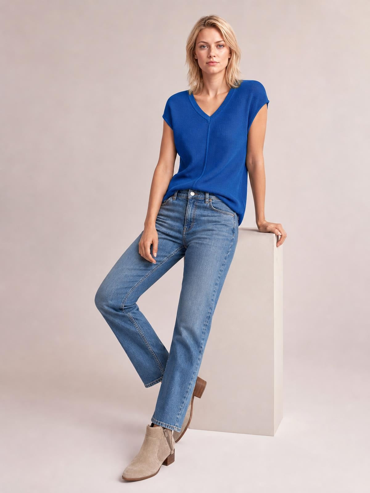A model wears a royal blue V-neck short sleeve knit top with a relaxed straight silhouette and textured construction, leaning against a cube in jeans.
