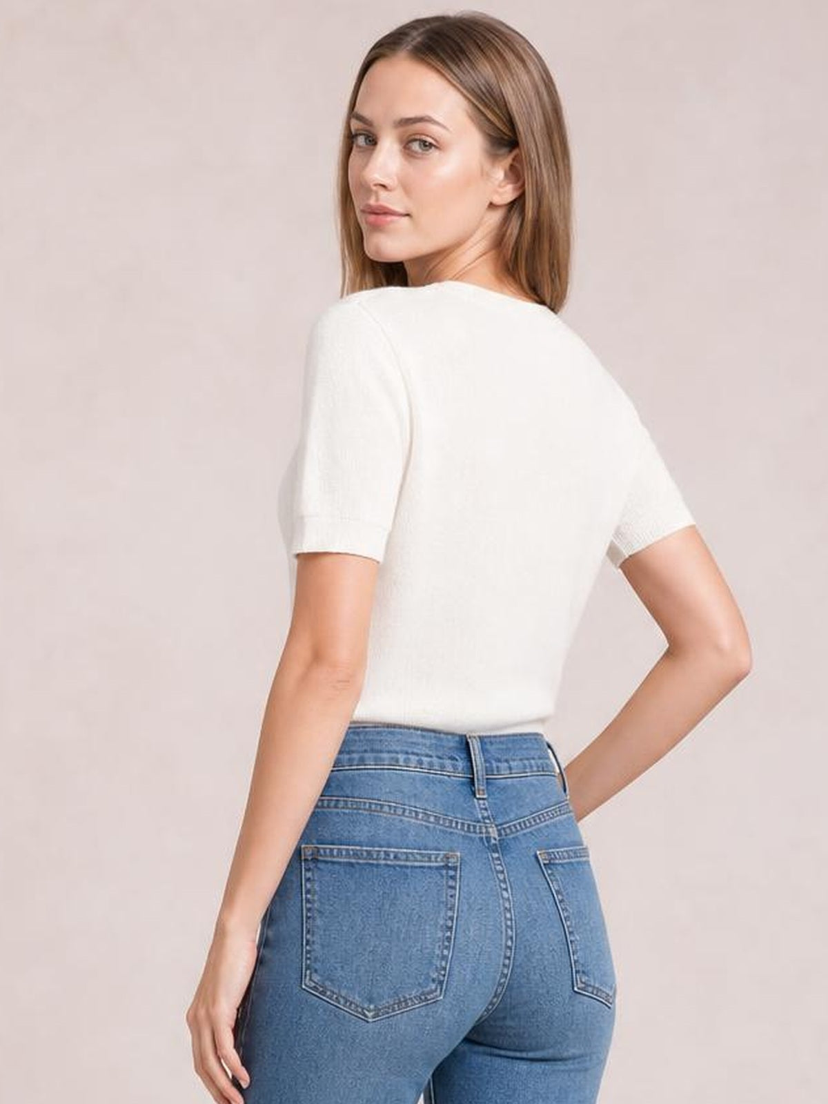 A model wears a white short-sleeve knit top with a relaxed straight silhouette, shown from the back while turning with one hand on her hip.