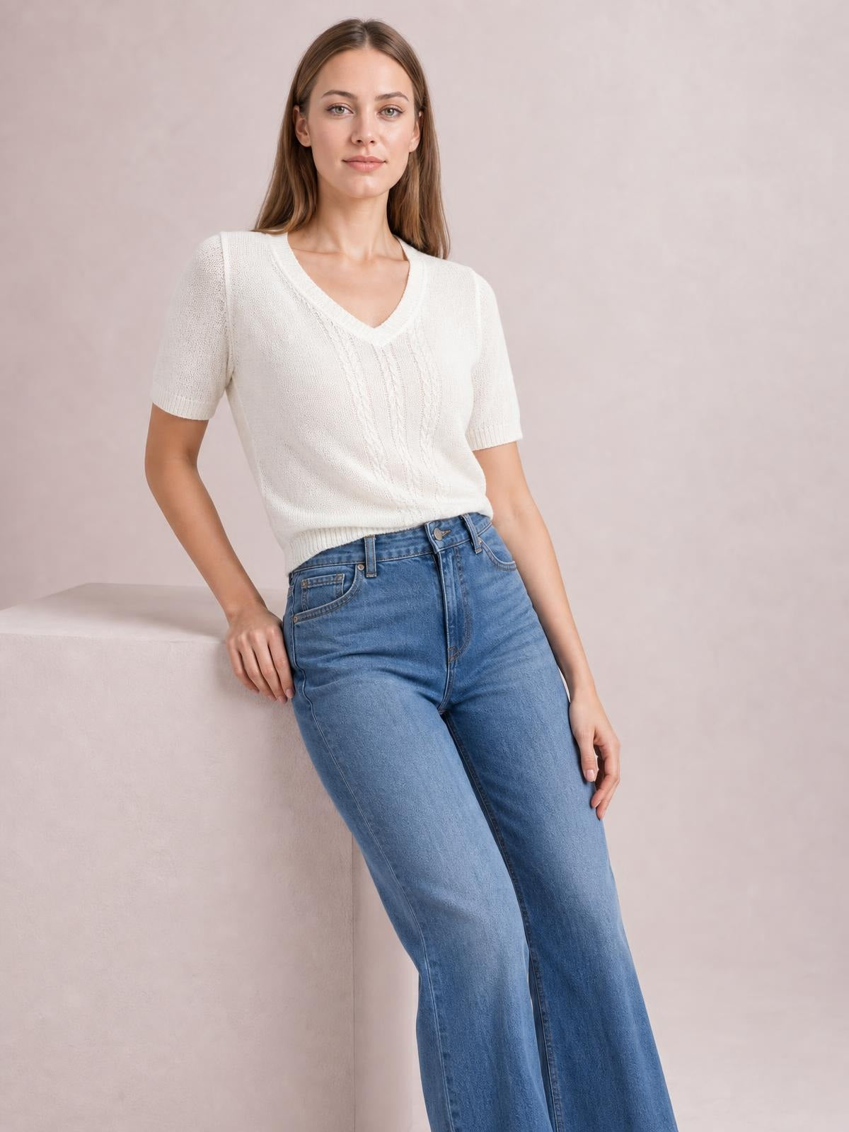 A model leans against a ledge wearing a white V-neck short sleeve knit top with a slim silhouette, ribbed trim, and subtle center cable-knit detail.