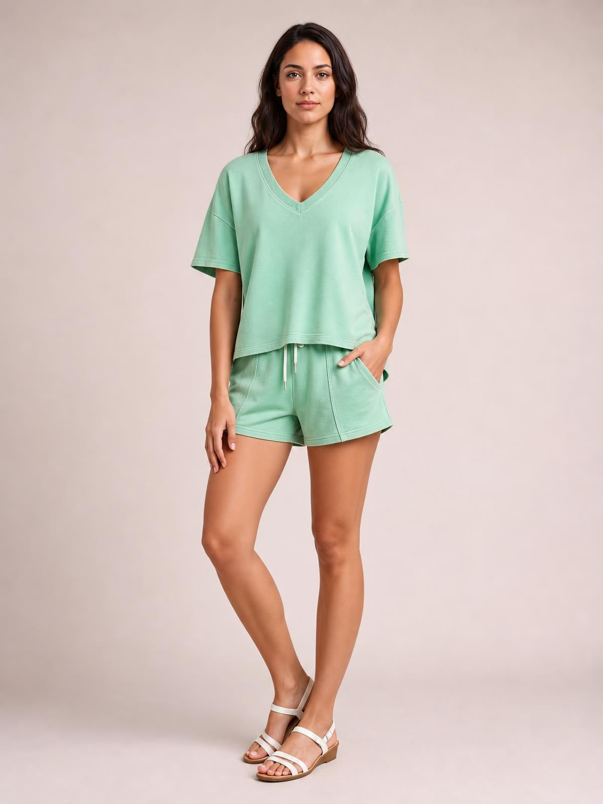 Woman poses in a mint green V-neck short sleeve loose top and drawstring shorts set with a relaxed silhouette, visible pockets, and slight stretch.