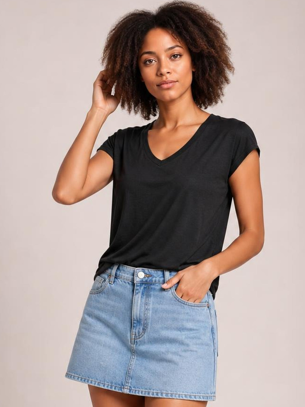 A model poses in a black V-neck short sleeve T-shirt with a relaxed straight silhouette, lightweight construction, and one hand resting in a denim skirt pocket.
