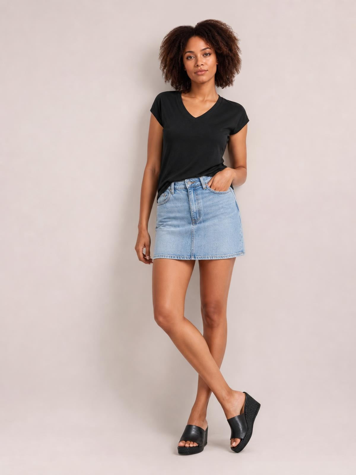 Model wearing a black V-neck short sleeve T-shirt with a basic straight silhouette, posing with one hand in her pocket and legs crossed.