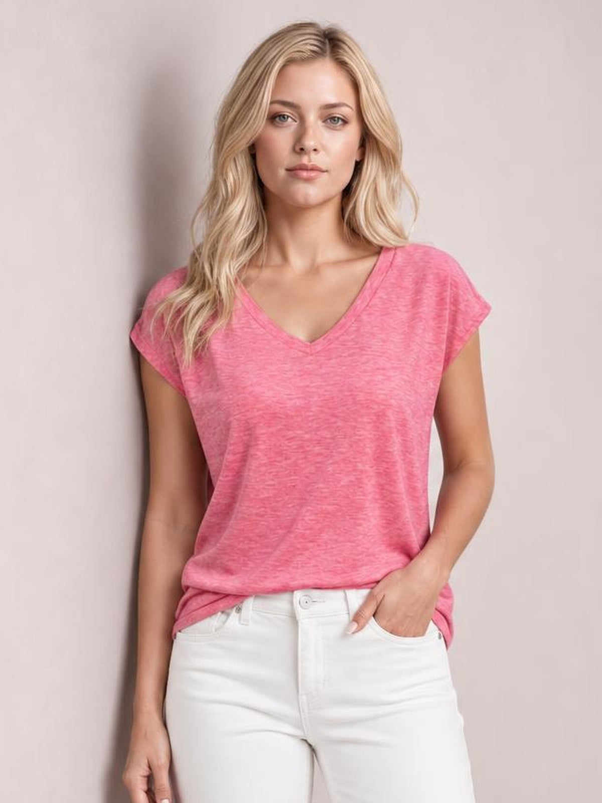 A model wears a pink V-neck short sleeve T-shirt with a relaxed, slightly loose silhouette, standing front-facing with one hand in her jeans pocket.