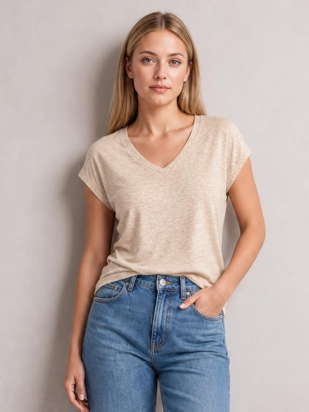 Woman wearing a beige V-neck short sleeve T-shirt with a relaxed straight silhouette, slightly dropped shoulders, and one hand in her jeans pocket.