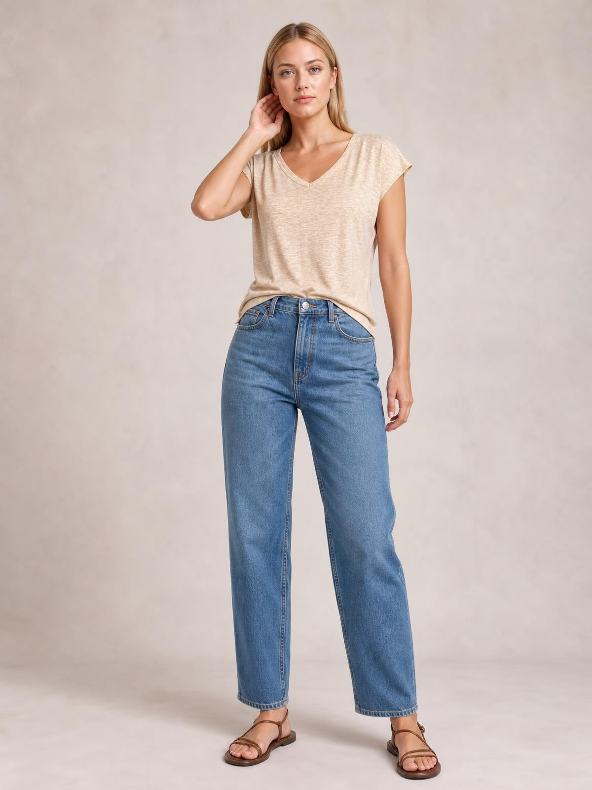 A model poses with one hand at her hair, wearing a beige V-neck short sleeve T-shirt with a relaxed silhouette and softly draped construction.