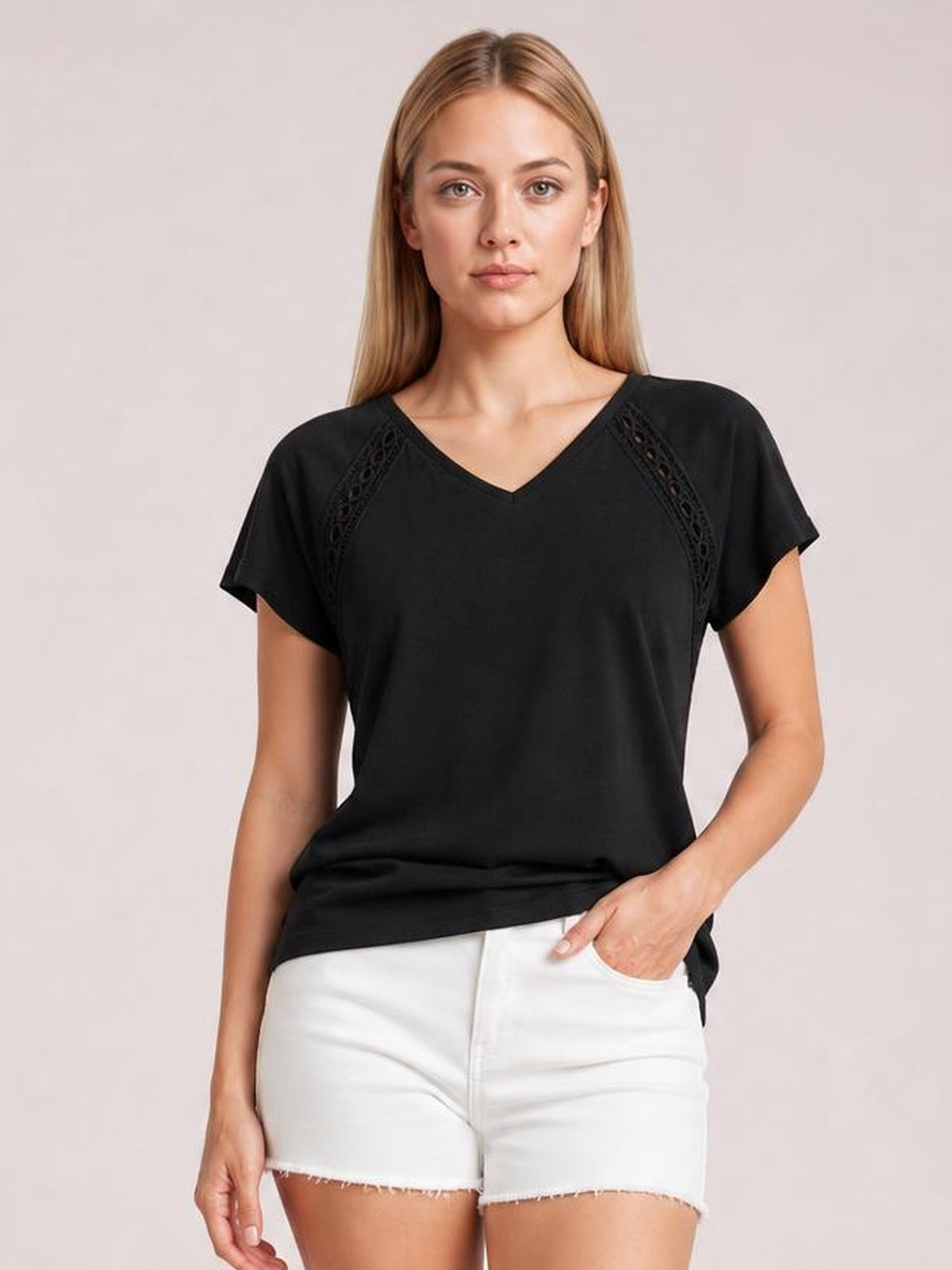 Model wearing a black V-neck short sleeve T-shirt with eyelet shoulder details and a relaxed silhouette, posing with one hand in her shorts pocket.