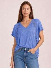 A model wears a blue V-neck short sleeve T-shirt with a relaxed silhouette and decorative eyelet panels, standing with one hand in her jeans pocket.