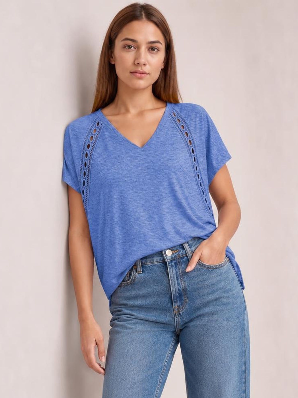 Model wearing a blue V-neck short sleeve T-shirt with a relaxed silhouette and decorative eyelet panels, posing with one hand in her jeans pocket.