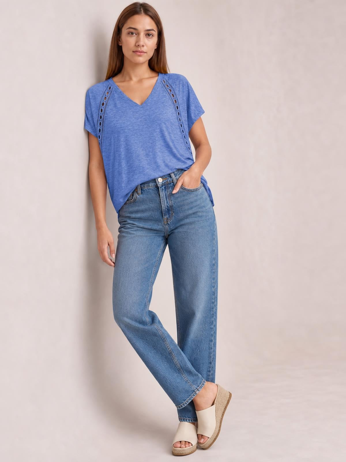 Model wears a blue V-neck short sleeve T-shirt with eyelet side details and a relaxed silhouette, posing with one hand in her pocket and crossed legs.