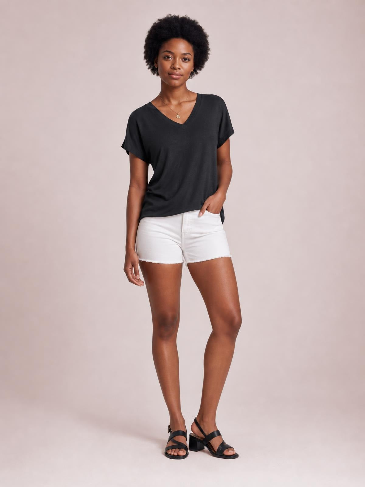 Model stands casually in a black V-neck short sleeve T-shirt with a relaxed straight silhouette, one hand resting in her white shorts pocket.