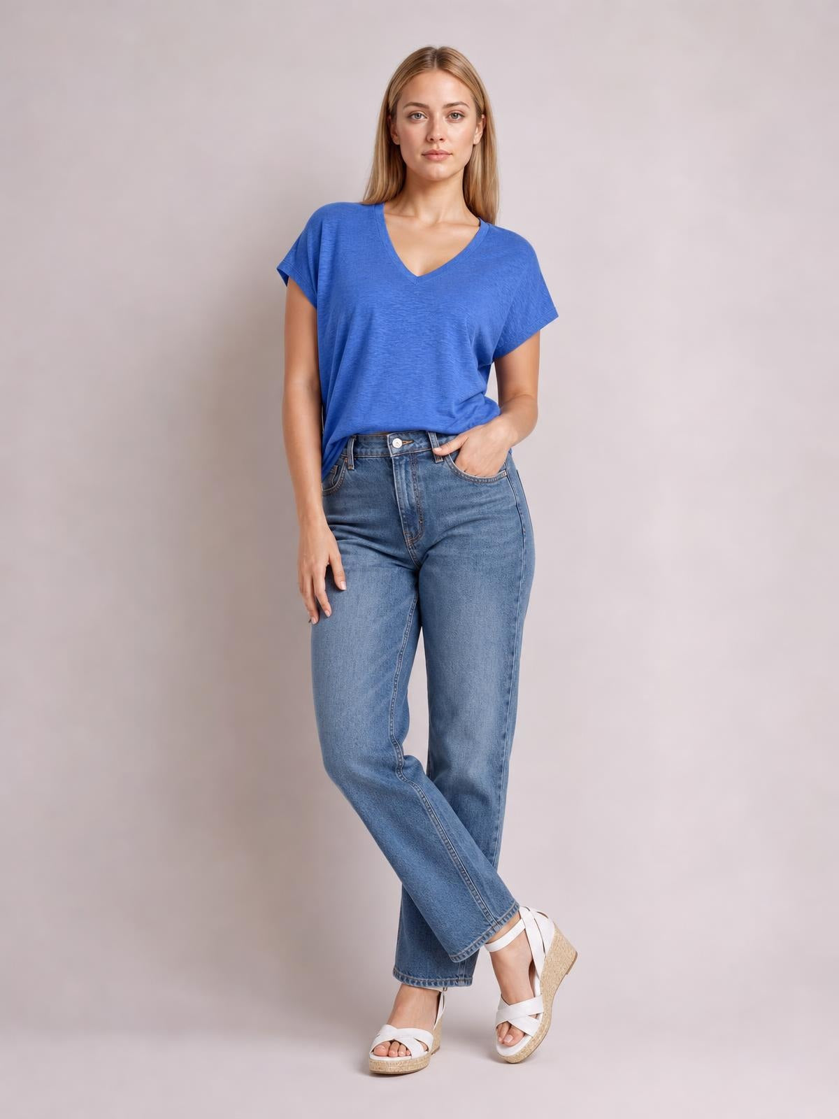 A woman models a blue V-neck short sleeve T-shirt with a relaxed silhouette, lightly tucked into jeans, standing with crossed legs and one hand in her pocket.