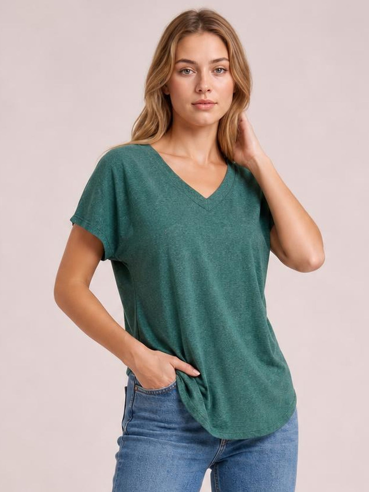 Model wearing a teal V-neck short sleeve T-shirt with a relaxed silhouette, curved hem, and slightly loose sleeves, posing with one hand in her jeans pocket.