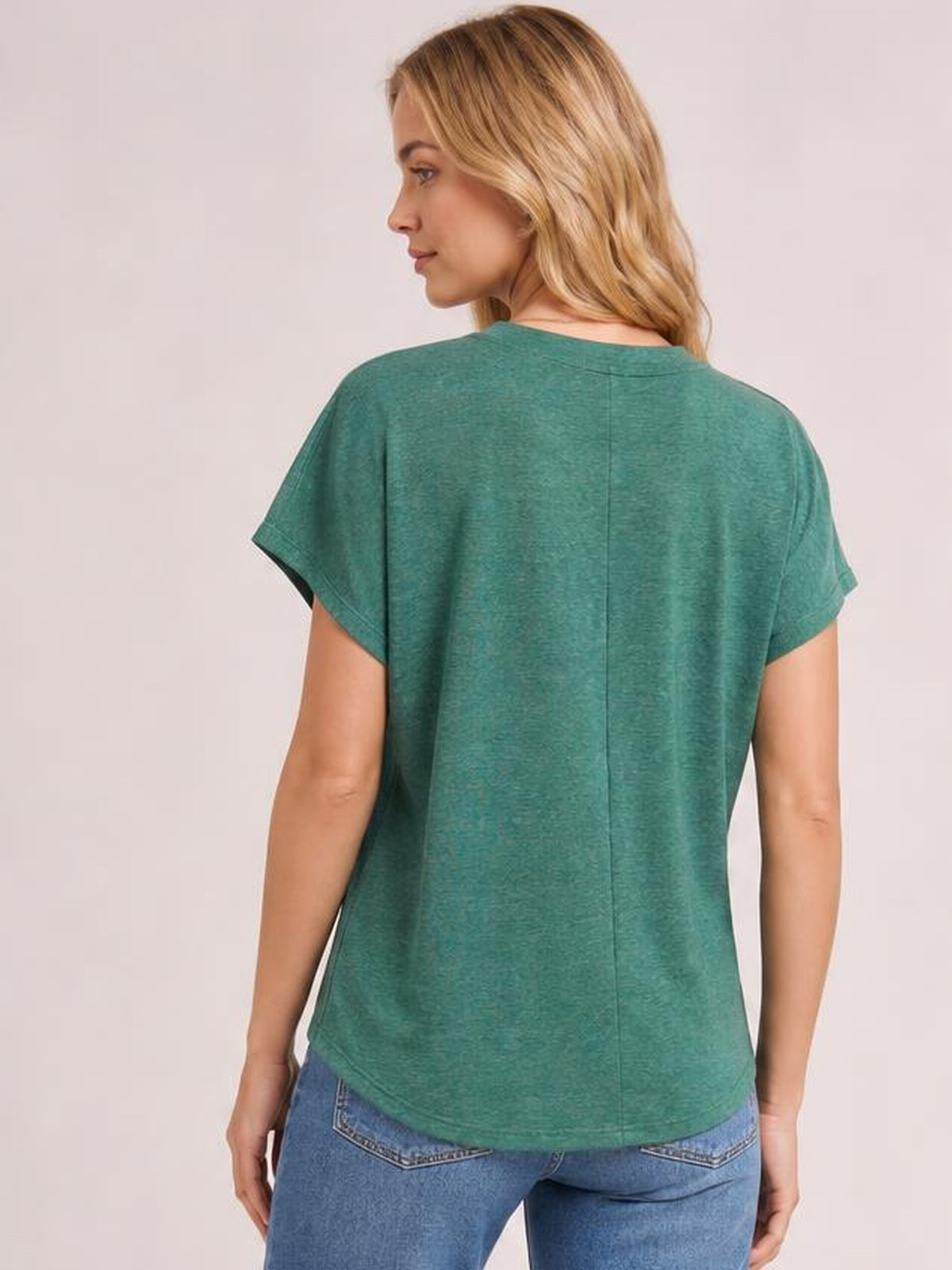 A model turned slightly to the side showcases the teal green short sleeve T-shirt’s relaxed silhouette, rounded hem, and center-back seam.