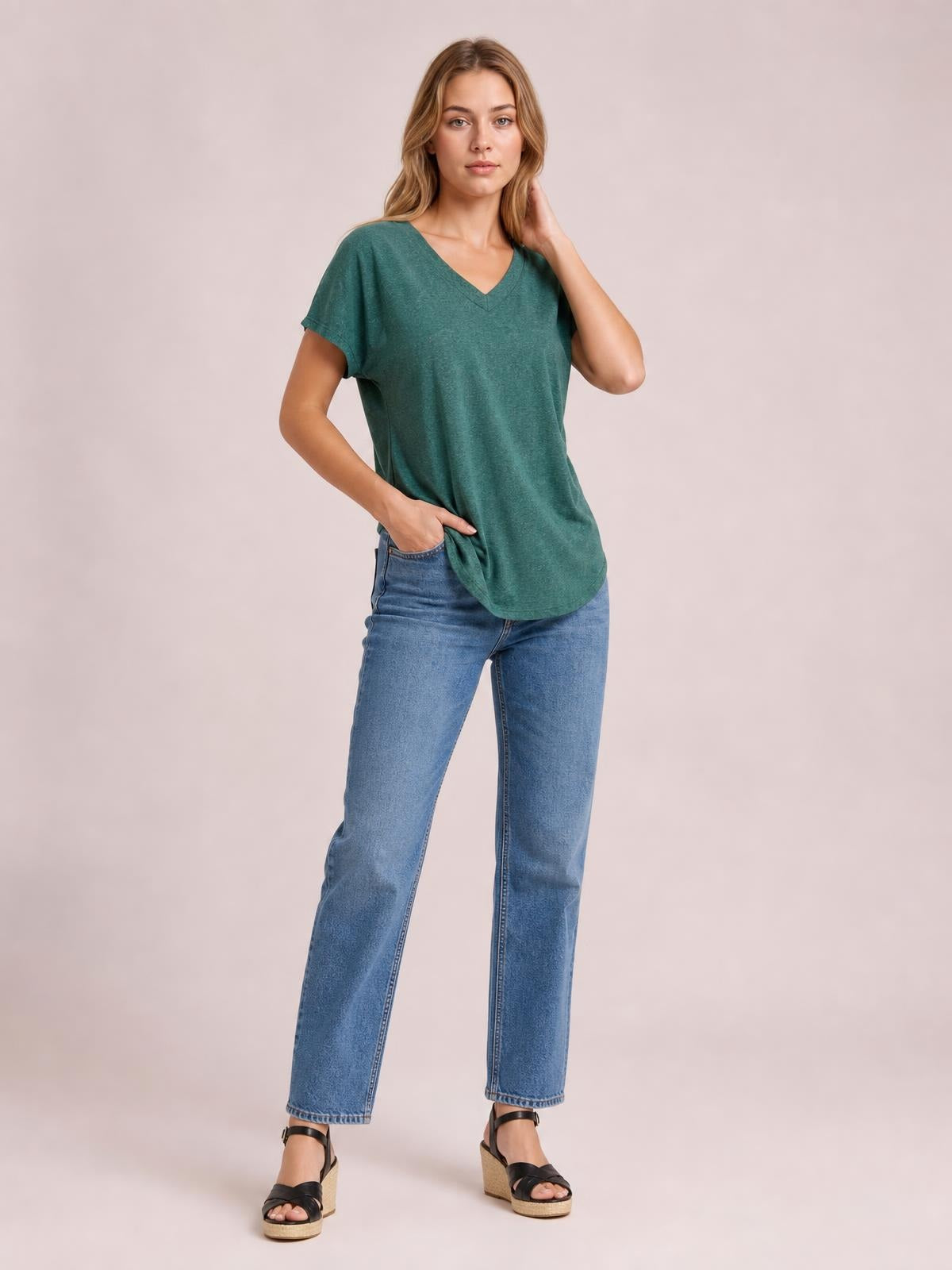 Model poses in a teal V-neck short sleeve T-shirt with a relaxed silhouette, curved hem, and slight stretch, paired with jeans.