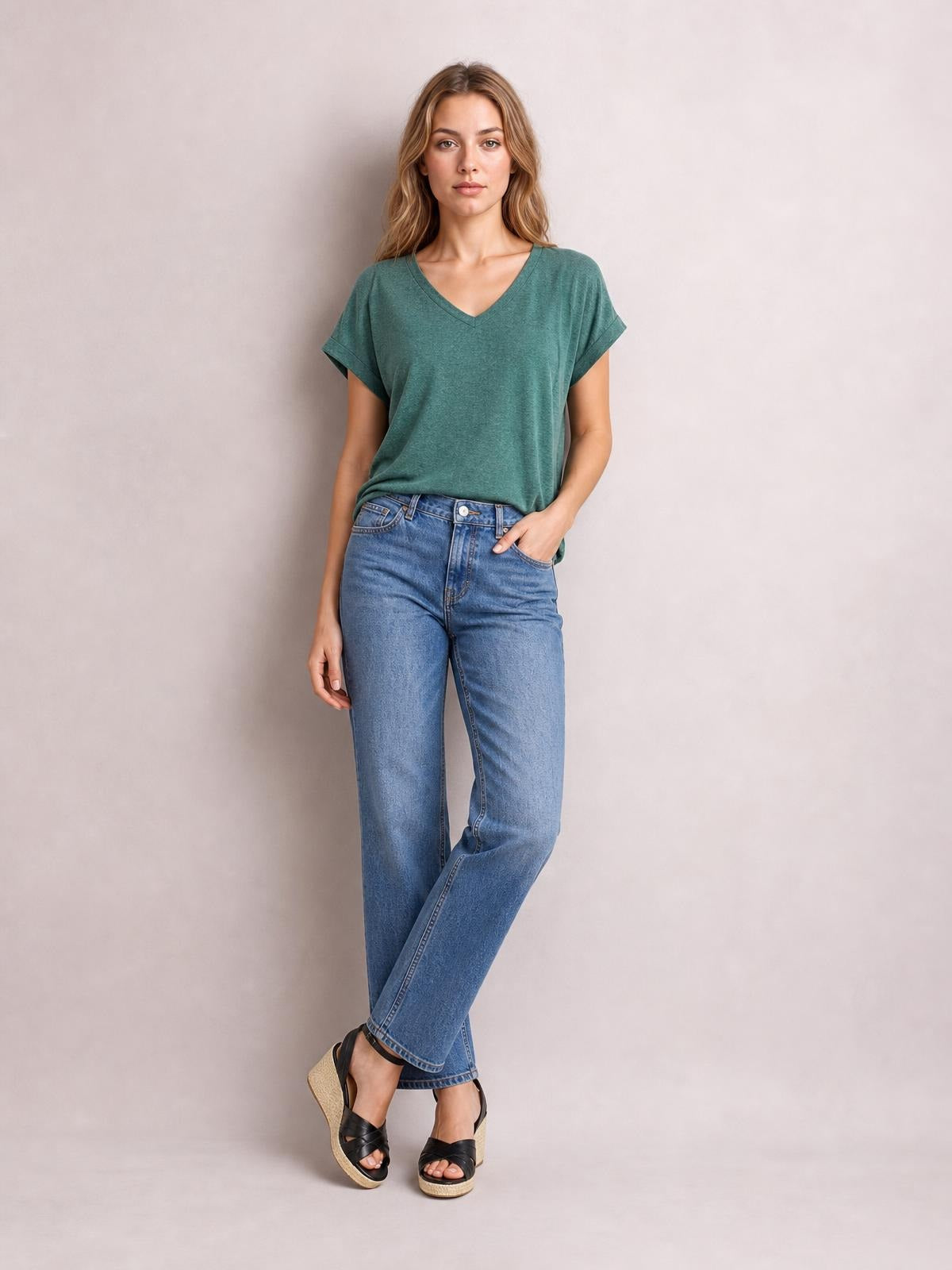 Model wearing a teal V-neck short sleeve T-shirt with a relaxed silhouette and rolled sleeve cuffs, casually posed with one hand in her jeans pocket.