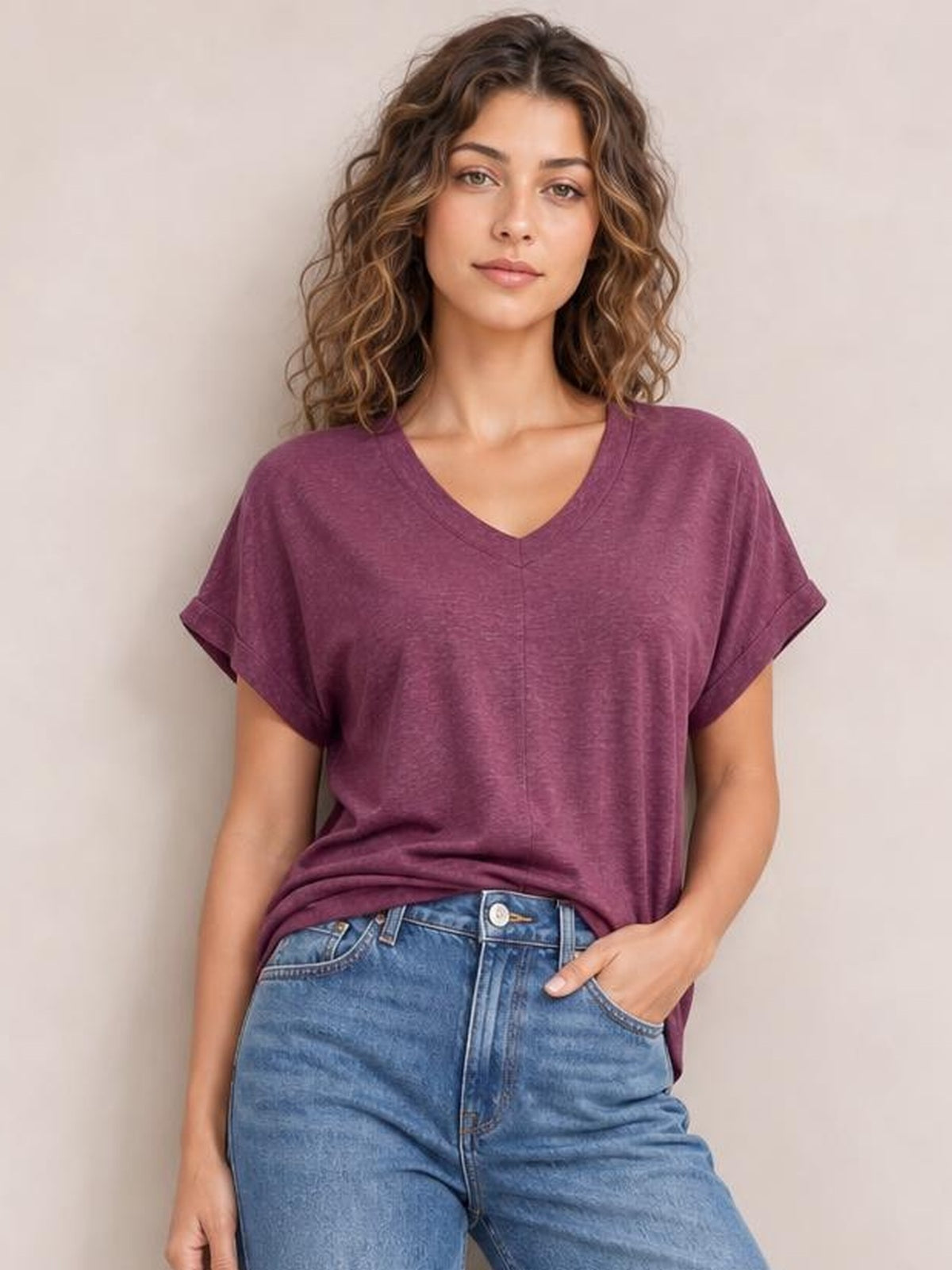 Model wearing a burgundy V-neck short sleeve T-shirt with cuffed sleeves, a relaxed silhouette, and a straight center seam, posed with one hand in her jeans pocket.