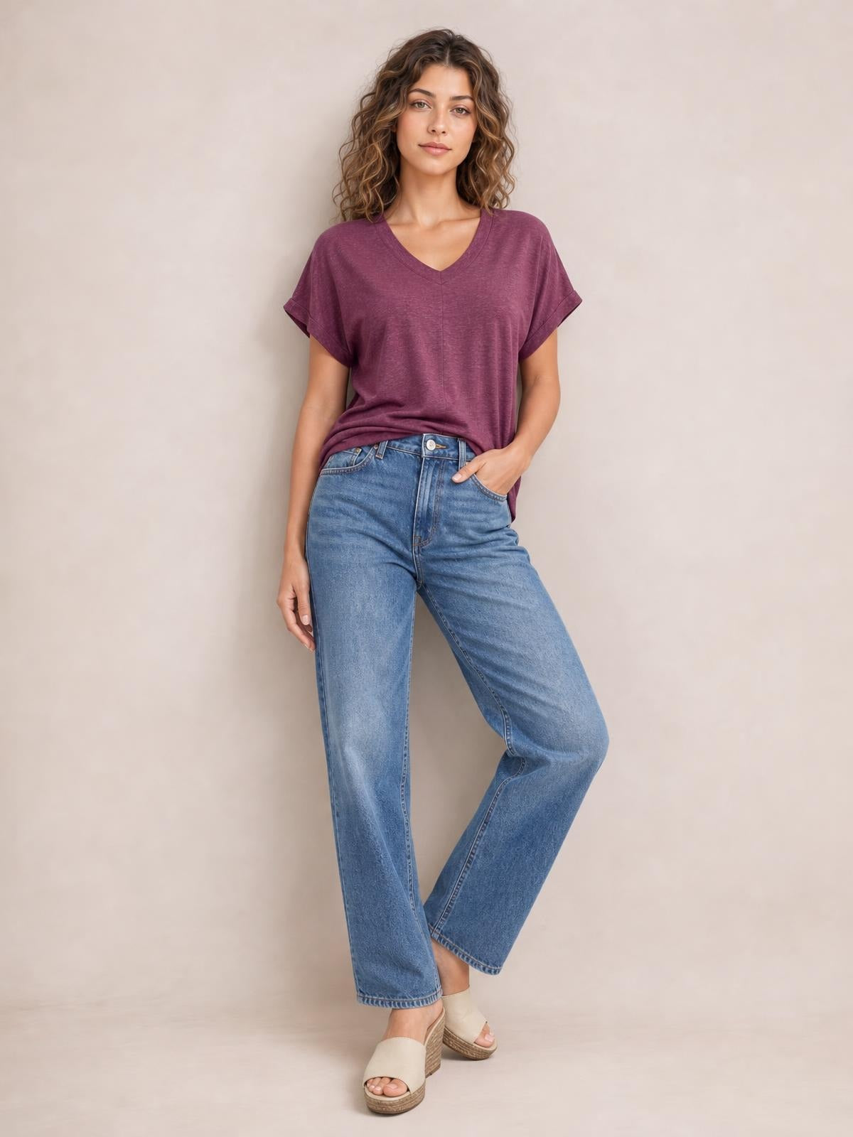 A model poses in a burgundy V-neck short sleeve T-shirt with a relaxed silhouette and rolled cuffs, paired with jeans and one hand in a pocket.