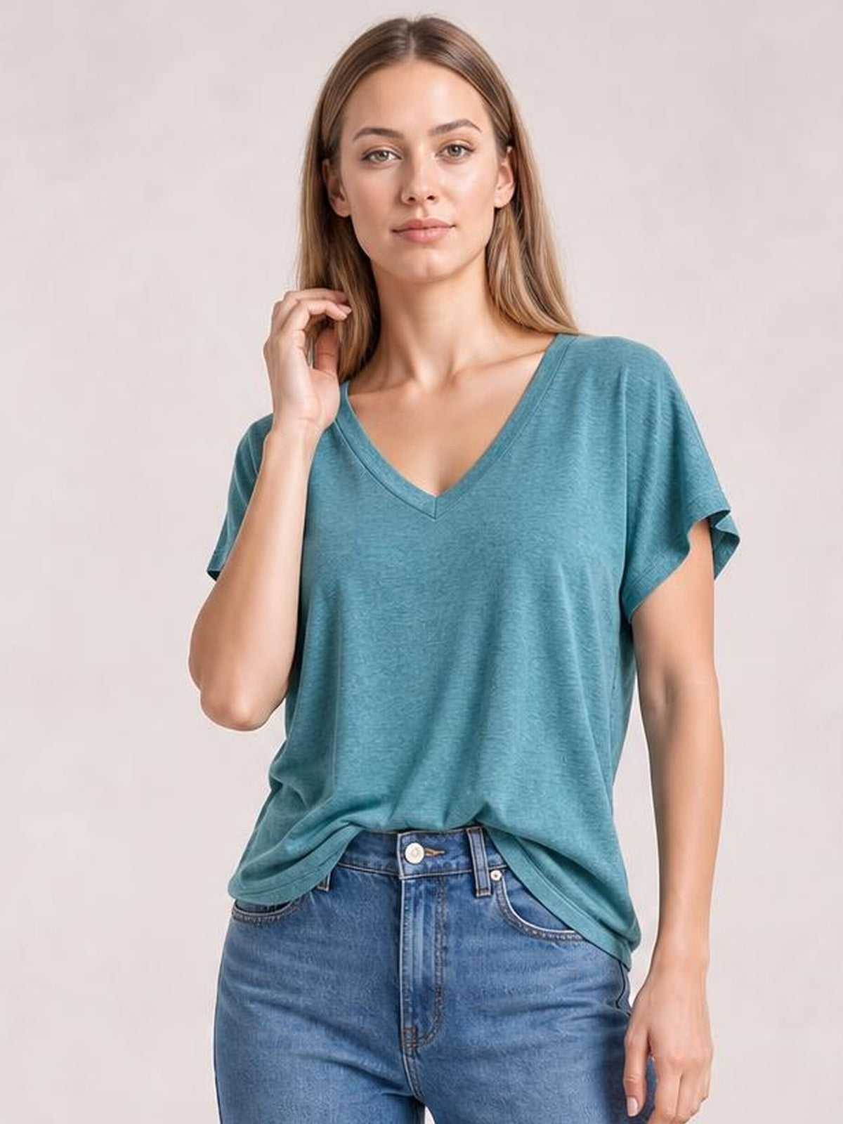 Model wearing a teal V-neck short sleeve T-shirt with a relaxed silhouette, slightly stretchy fabric, and rolled sleeve cuffs, posing with one hand near her hair.