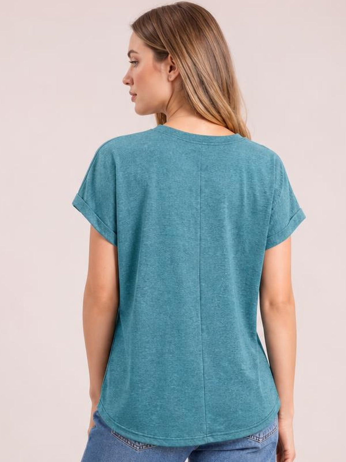 A model looks over her shoulder in a teal short sleeve T-shirt with rolled cuffs, a relaxed silhouette, rounded hem, and visible center-back seam.