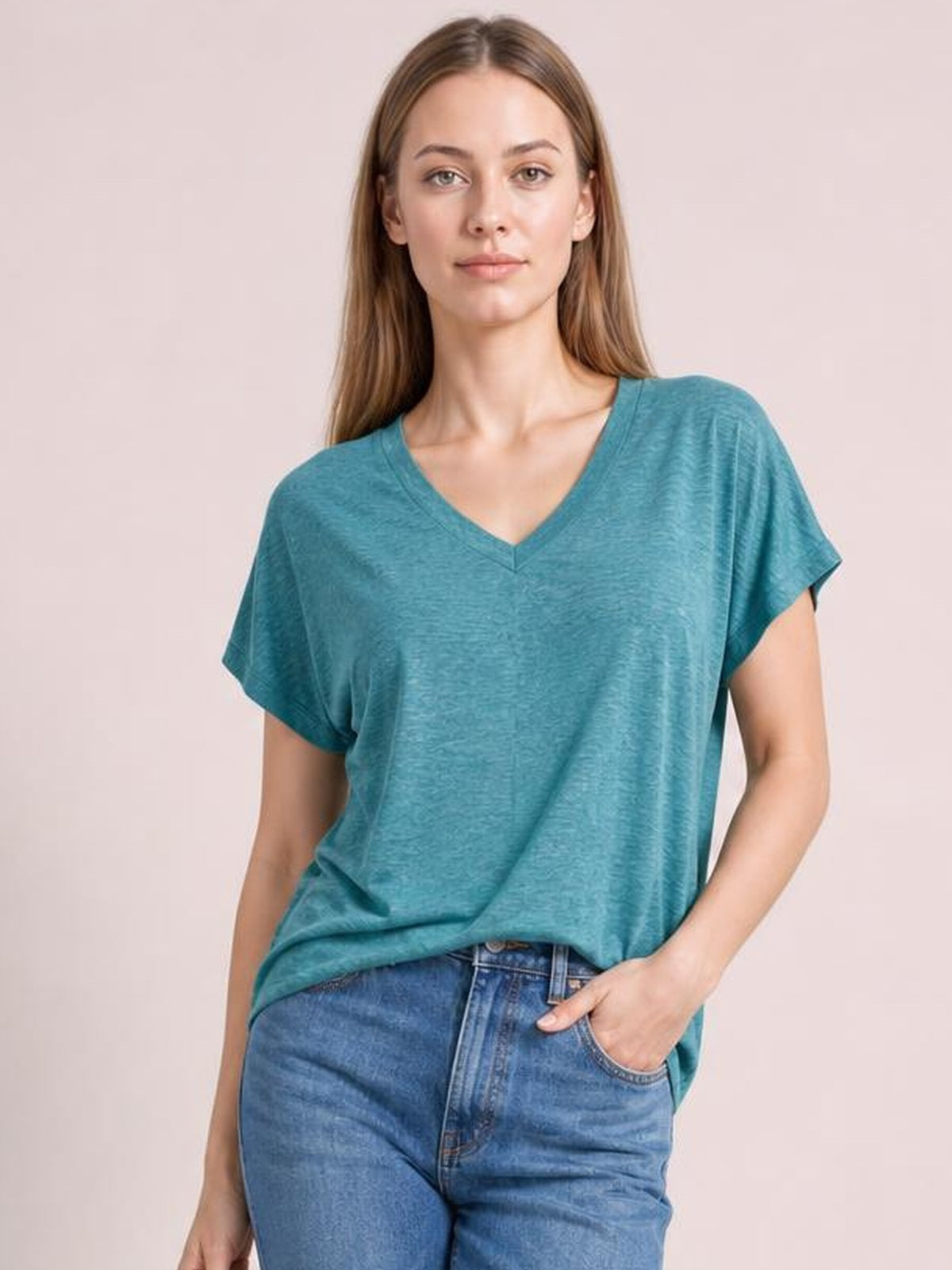 Model wearing a teal V-neck short sleeve T-shirt with a relaxed silhouette, draped fit, and slightly rolled sleeve hems, posing with one hand in her jeans pocket.