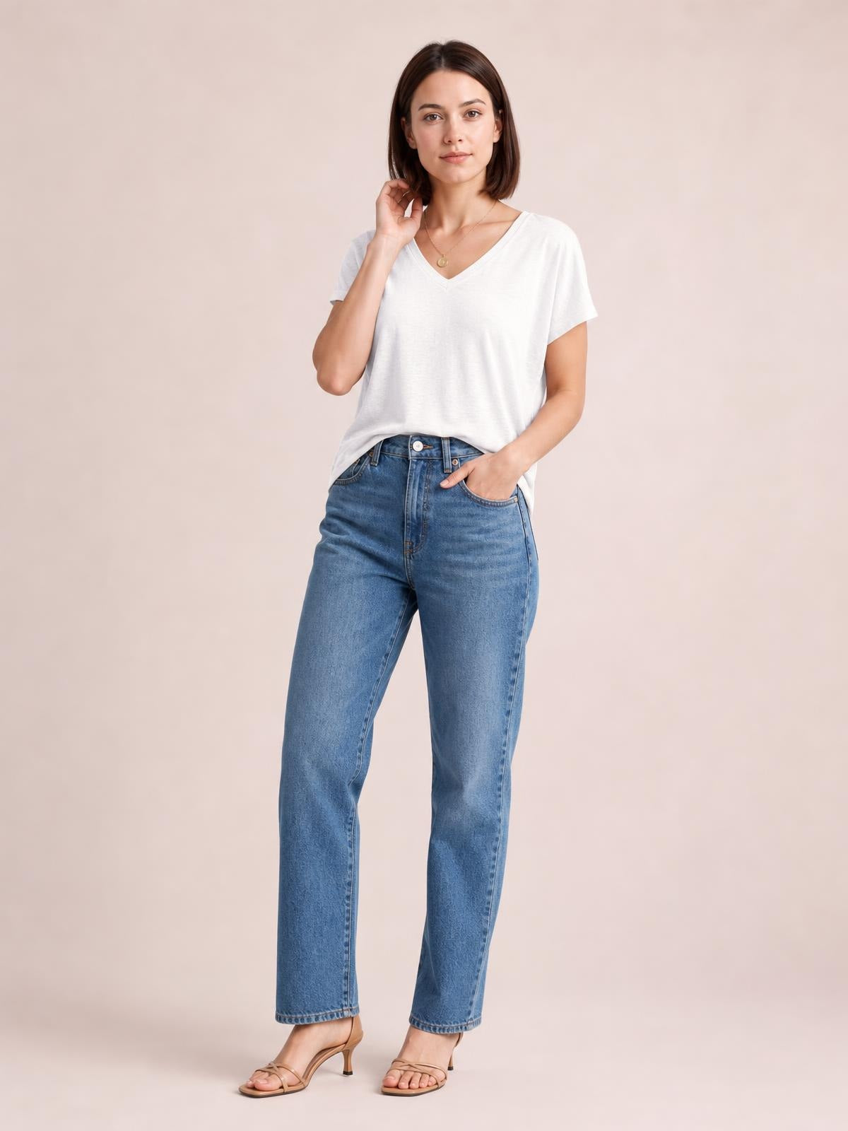 A model poses in a white V-neck short sleeve T-shirt with a relaxed, slightly loose silhouette, one hand touching her hair and the other in a jeans pocket.