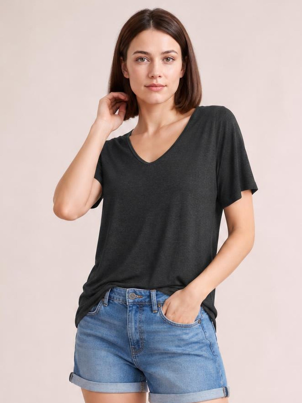 Model wearing a charcoal V-neck short sleeve T-shirt with a relaxed straight silhouette and finished neckline, one hand touching her hair and the other in her pocket.
