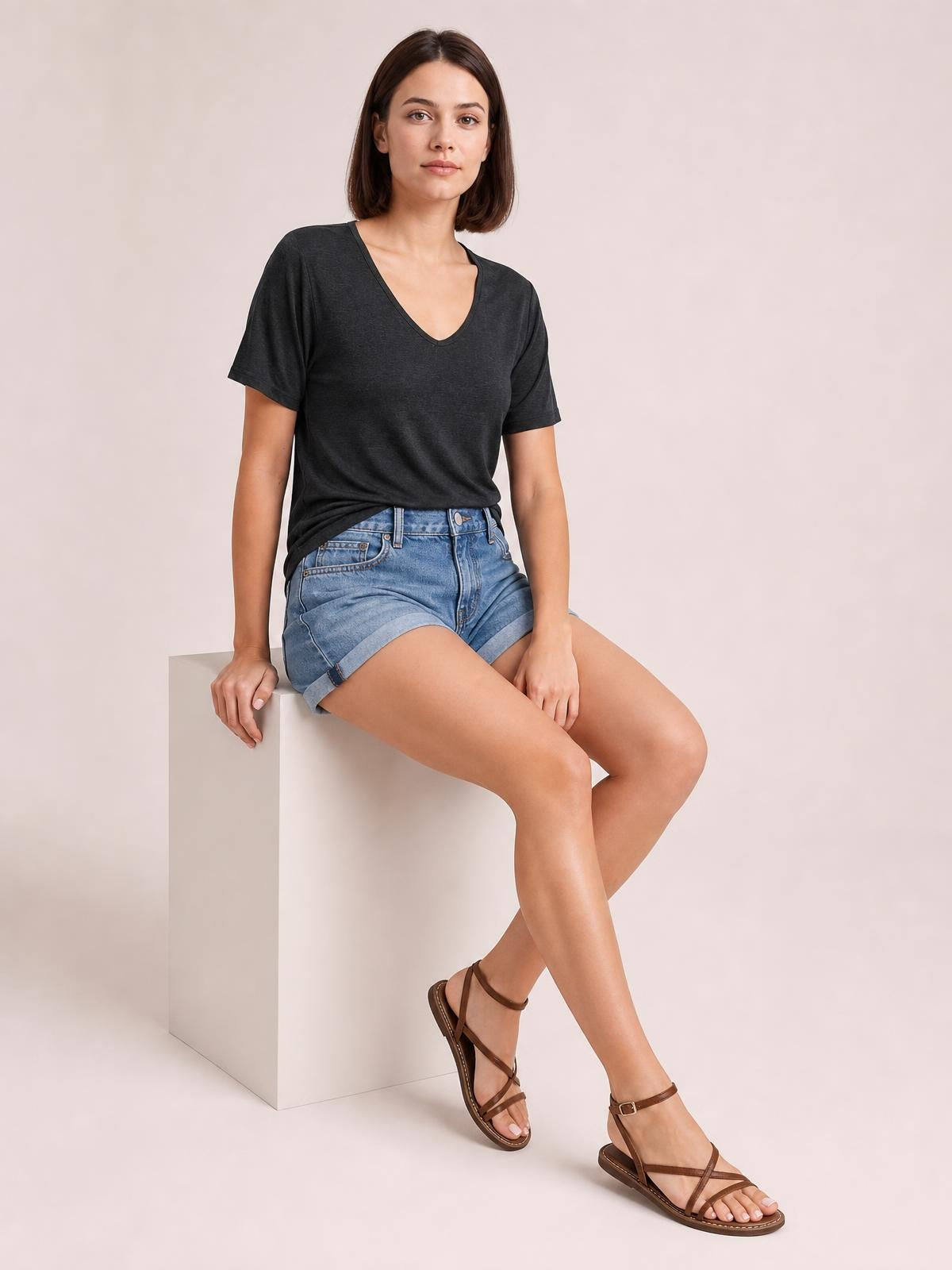 A woman seated in a relaxed pose wears a charcoal V-neck short sleeve T-shirt with a simple straight silhouette and lightly draped construction.