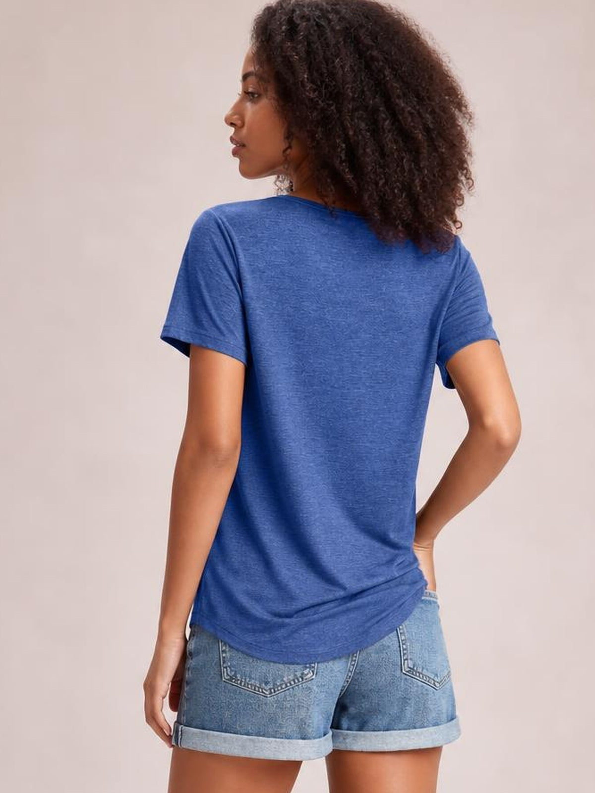 A model wears a blue short-sleeve T-shirt in a relaxed straight silhouette, shown from the back with a curved hem and one hand resting on her hip.