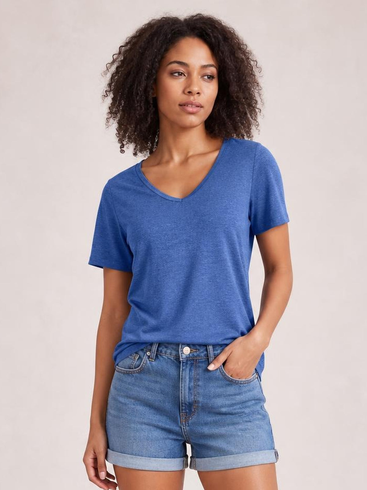 A model stands in a blue V-neck short sleeve T-shirt with a relaxed silhouette, slightly stretchy fabric, and one hand tucked into denim shorts.