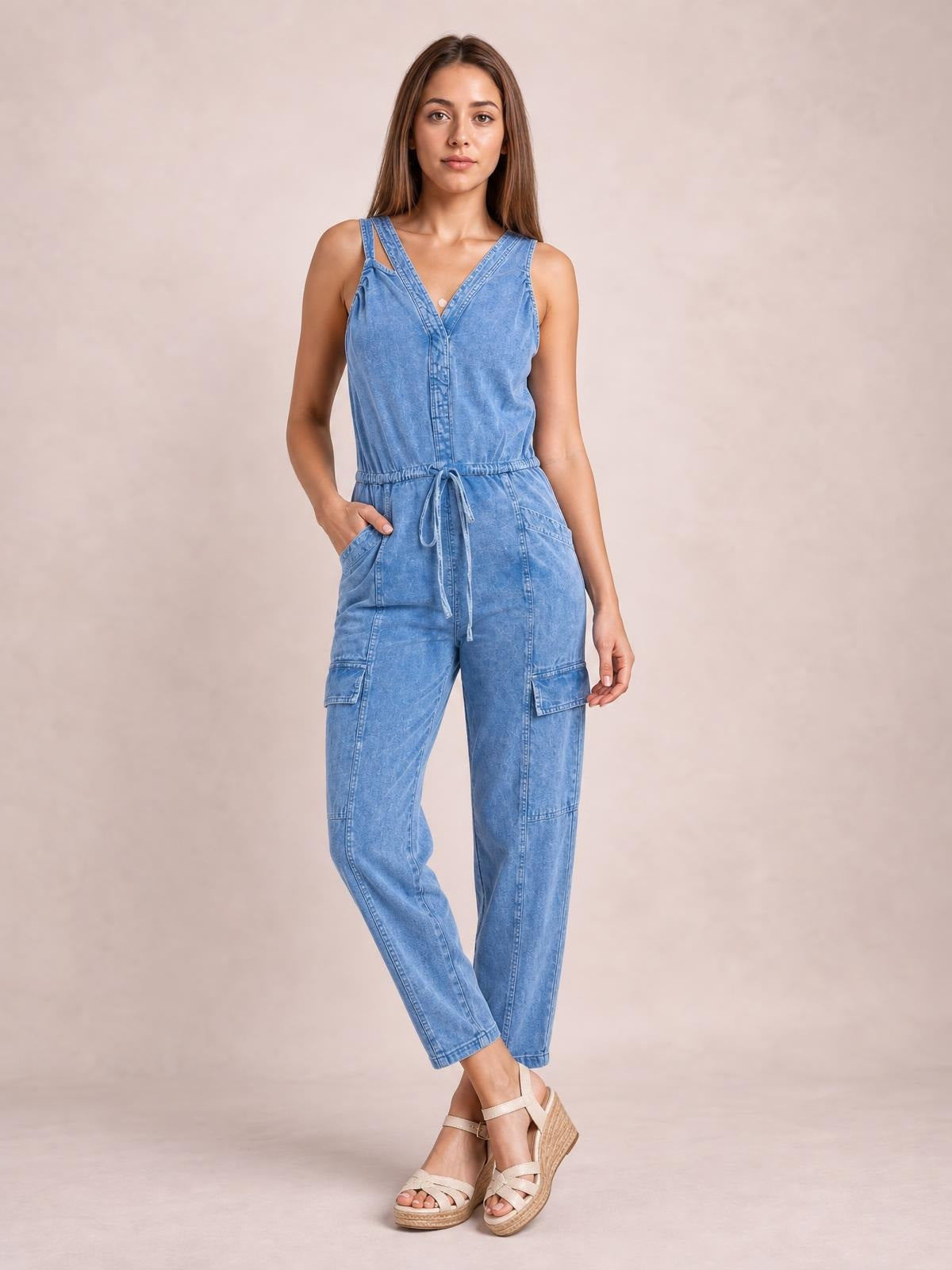 Model poses in a light blue sleeveless denim jumpsuit with a V-neck, adjustable drawstring waist, cargo-style pockets, and a relaxed tapered-leg silhouette.