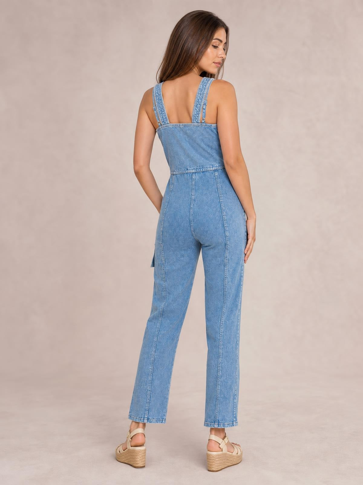 Model poses from the back in a light blue sleeveless denim jumpsuit with shoulder straps, a defined waist, straight-leg silhouette, and visible seam detailing.